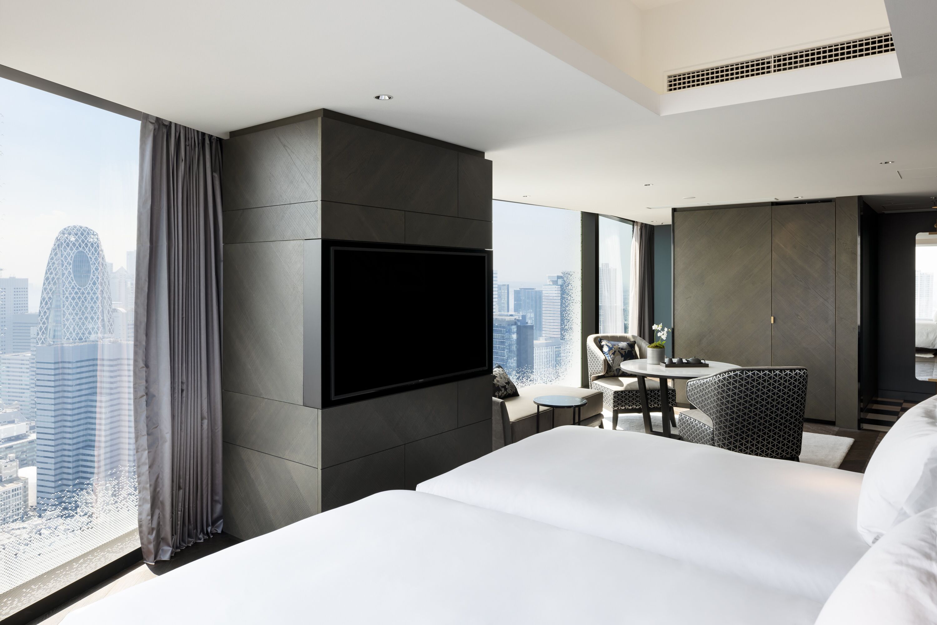 A modern hotel room with a large bed, a flat-screen TV mounted on the wall, a small round table with chairs, and a view of the cityscape through large windows.
