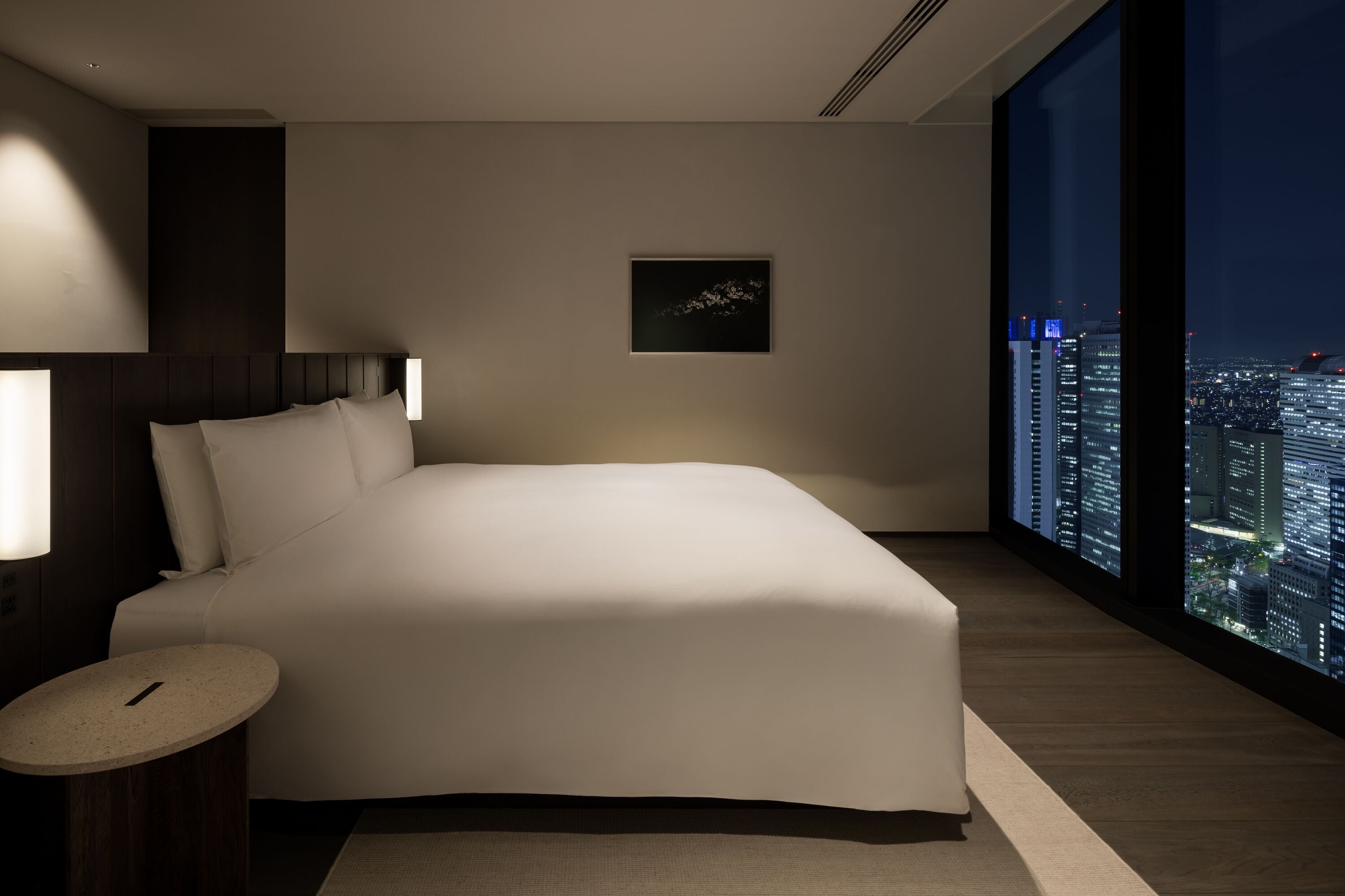 A modern hotel room with a large bed, a small round table, a framed artwork on the wall, and a city view through large windows.