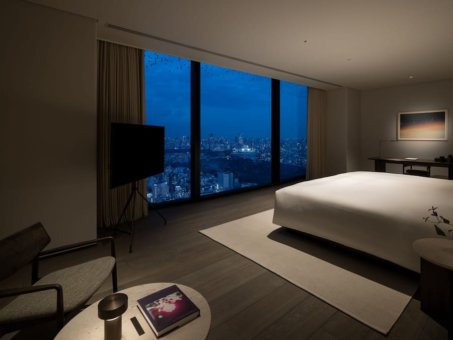 A modern hotel room with a large window offering a city view, a bed, a desk, and a television.