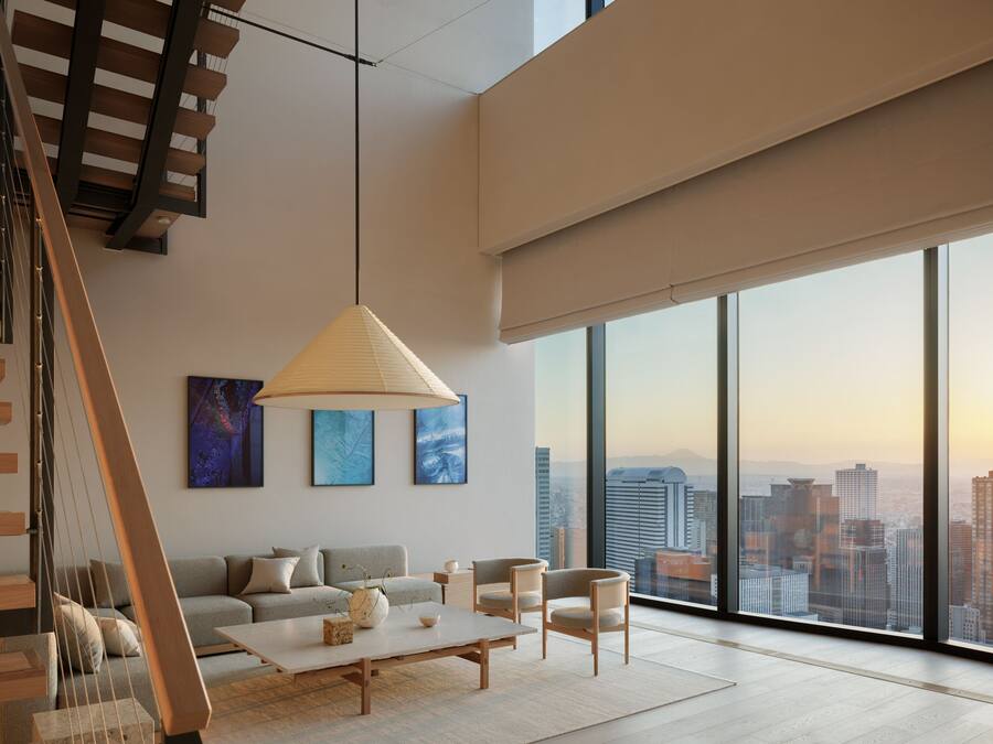 A spacious living area with a high ceiling, a staircase, and a large window offering a city view.