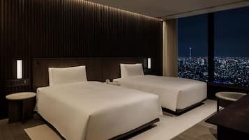 A modern hotel room with two beds, a nightstand, a desk, and a large window offering a city view.