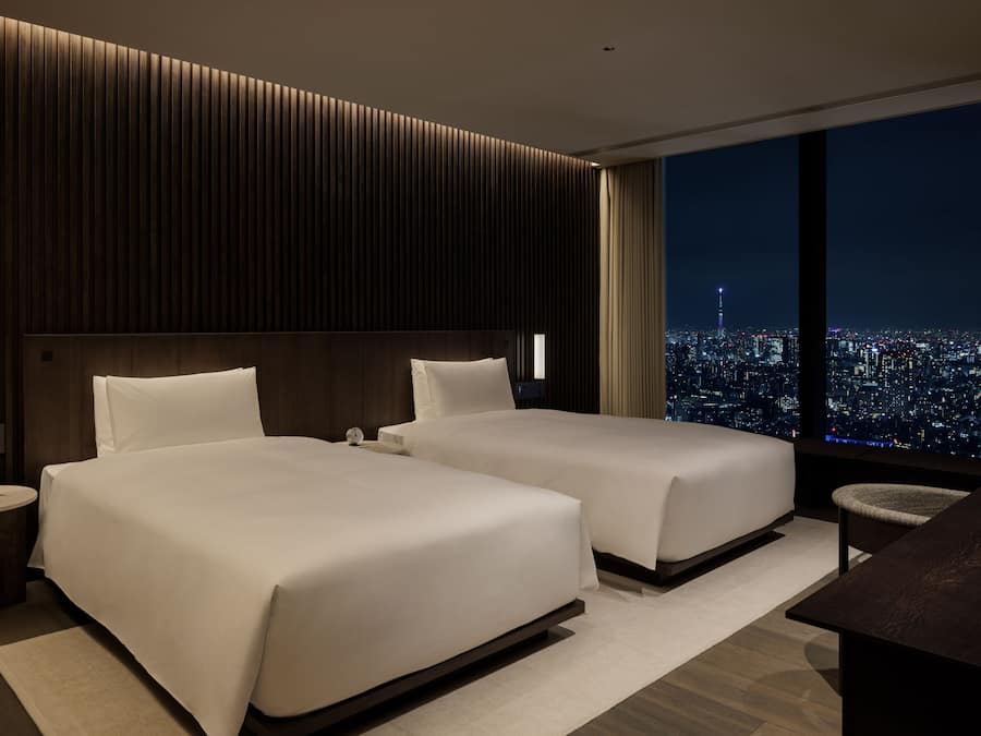 A modern hotel room with two beds, a nightstand, a desk, and a large window offering a city view.