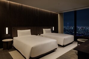 A modern hotel room with two beds, a nightstand, a desk, and a large window offering a city view.