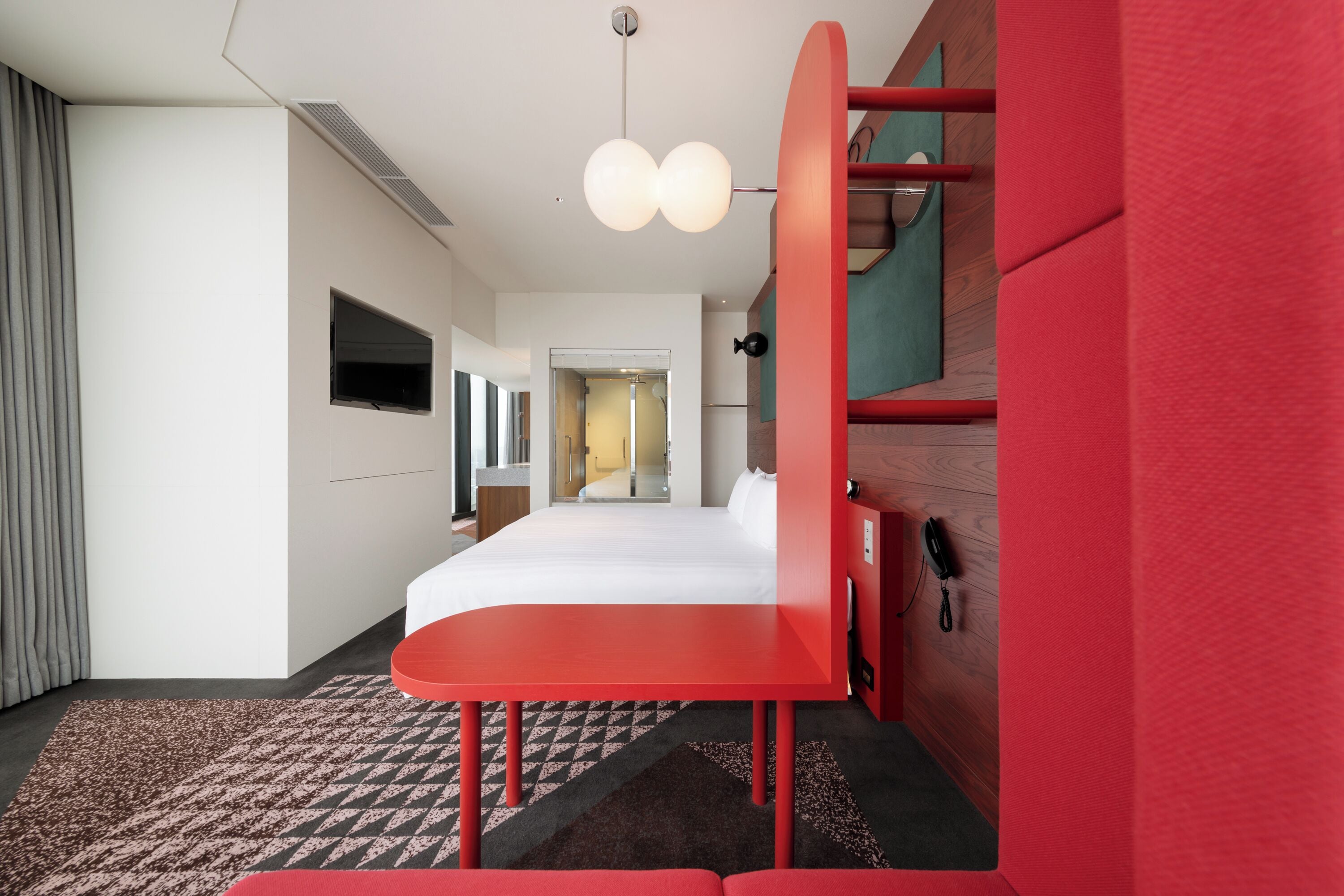 A hotel room with a bed, a red desk, a telephone, and a television on the wall.