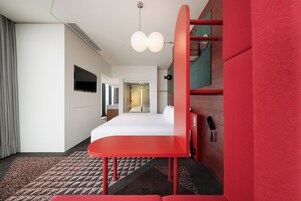 A hotel room with a bed, a red desk, a telephone, and a television on the wall.