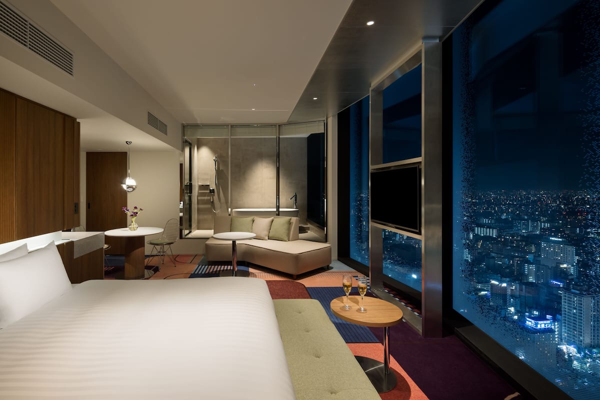 Ett modernt hotellrum med en stor säng, en sittgrupp med soffa och fåtöljer, ett litet bord med drycker och ett stort fönster med utsikt över staden.