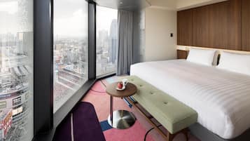 A hotel room with a large bed, a small round table with a cup, a bench, and a view of a cityscape through large windows.