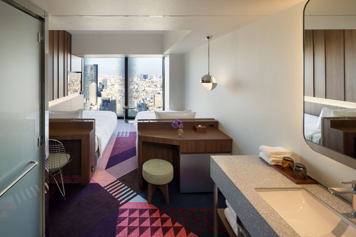 Ett hotellrum med en säng, ett skrivbord, ett badrum med handfat och ett stort fönster med utsikt över staden.