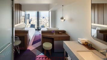 A hotel room with a bed, a desk, a bathroom with a sink, and a large window offering a city view.