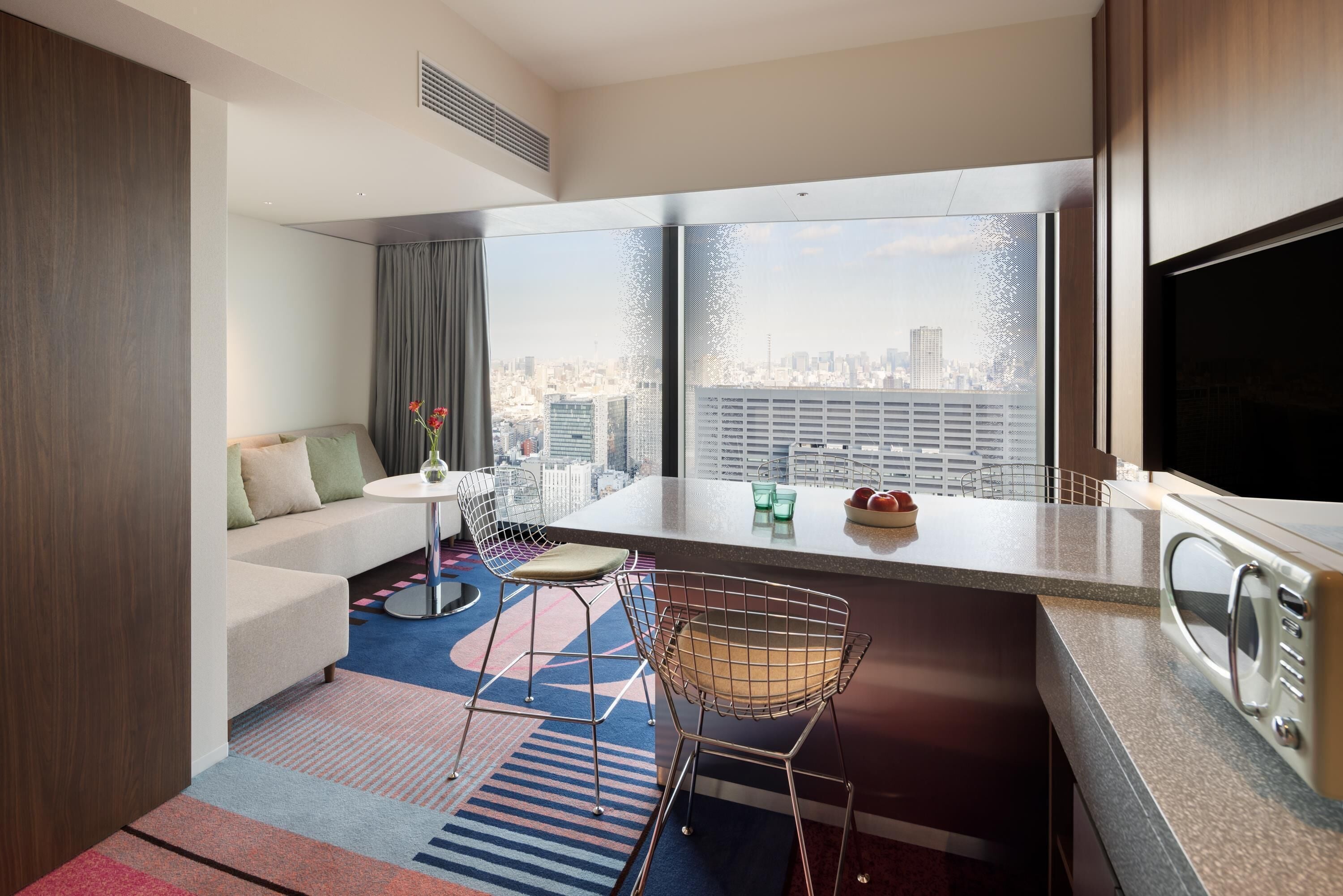 A modern hotel room with a city view, a kitchenette, a dining area, and a seating space.