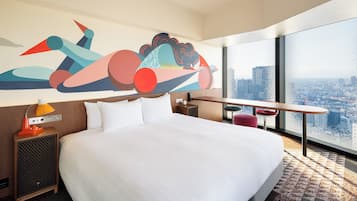 A hotel room with a large bed, a desk, a chair, a city view, and a colorful abstract mural.