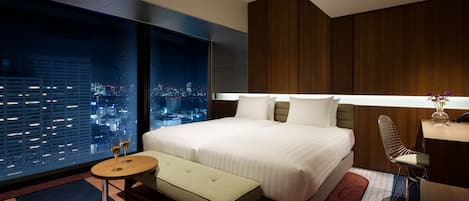 A modern hotel room with a large bed, a small round table, a bench, a desk, and a view of a cityscape at night.