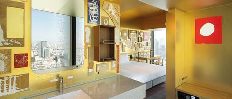 Habitación de hotel con un espejo grande, una cama, tocador con lavamanos y vistas a la ciudad.