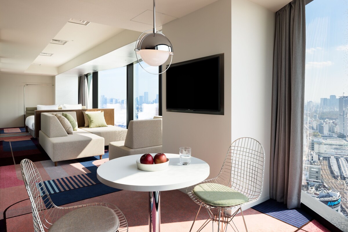 A modern hotel room with a city view, a round dining table, two chairs, a sofa, and a flat-screen TV.