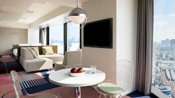 A modern hotel room with a city view, a round dining table, two chairs, a sofa, and a flat-screen TV.