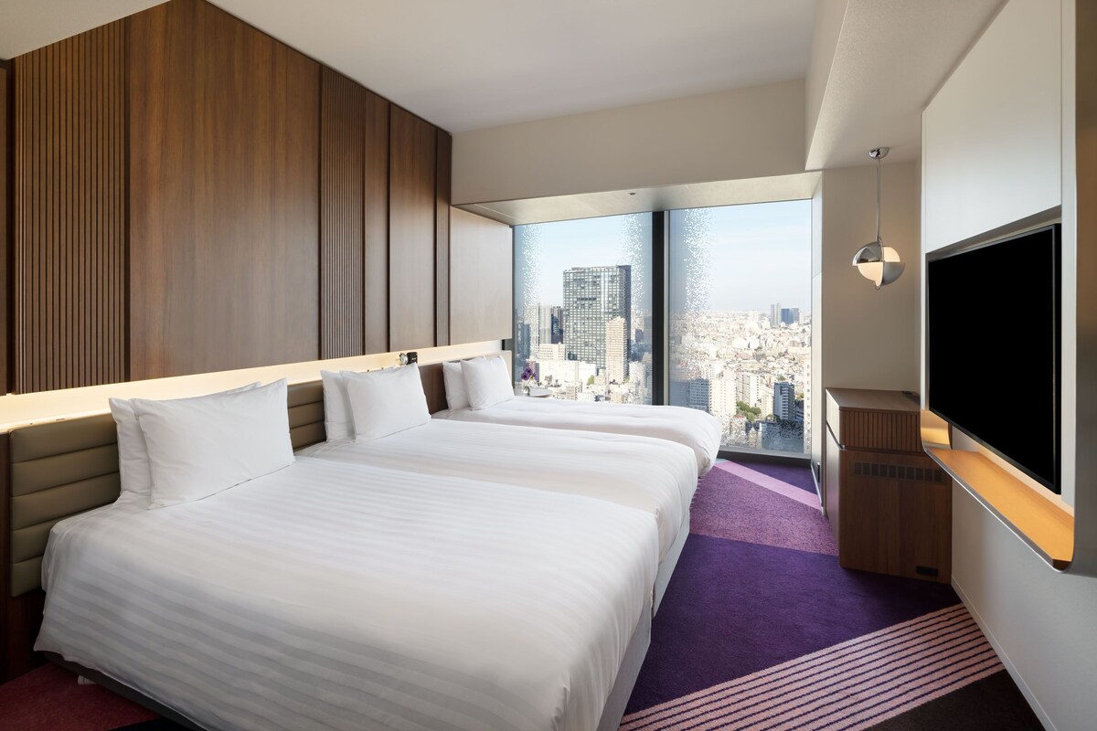 A hotel room with two beds, a large window with a city view, and a flat-screen TV.