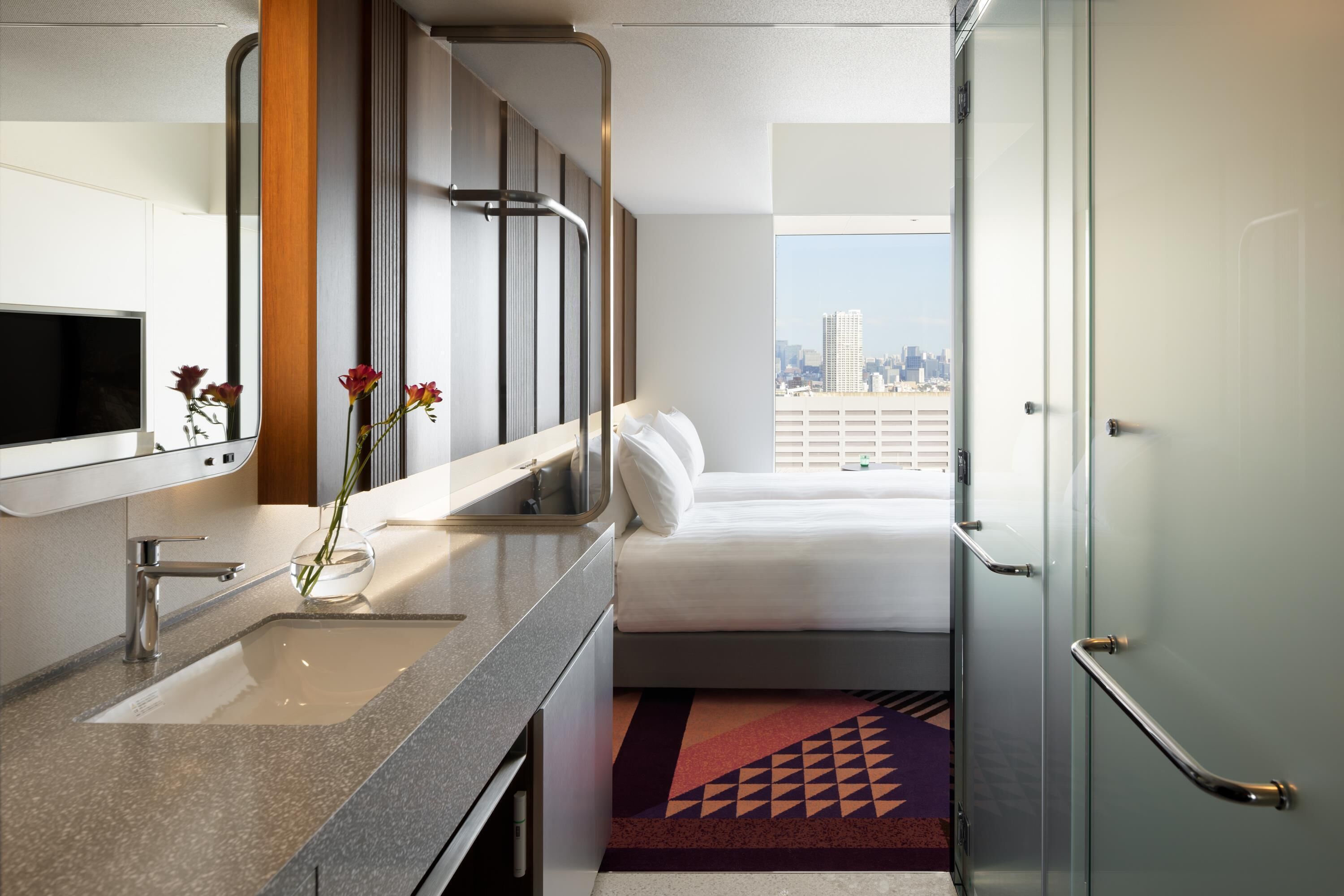 A hotel room with a bed, a bathroom with a sink and mirror, and a city view.