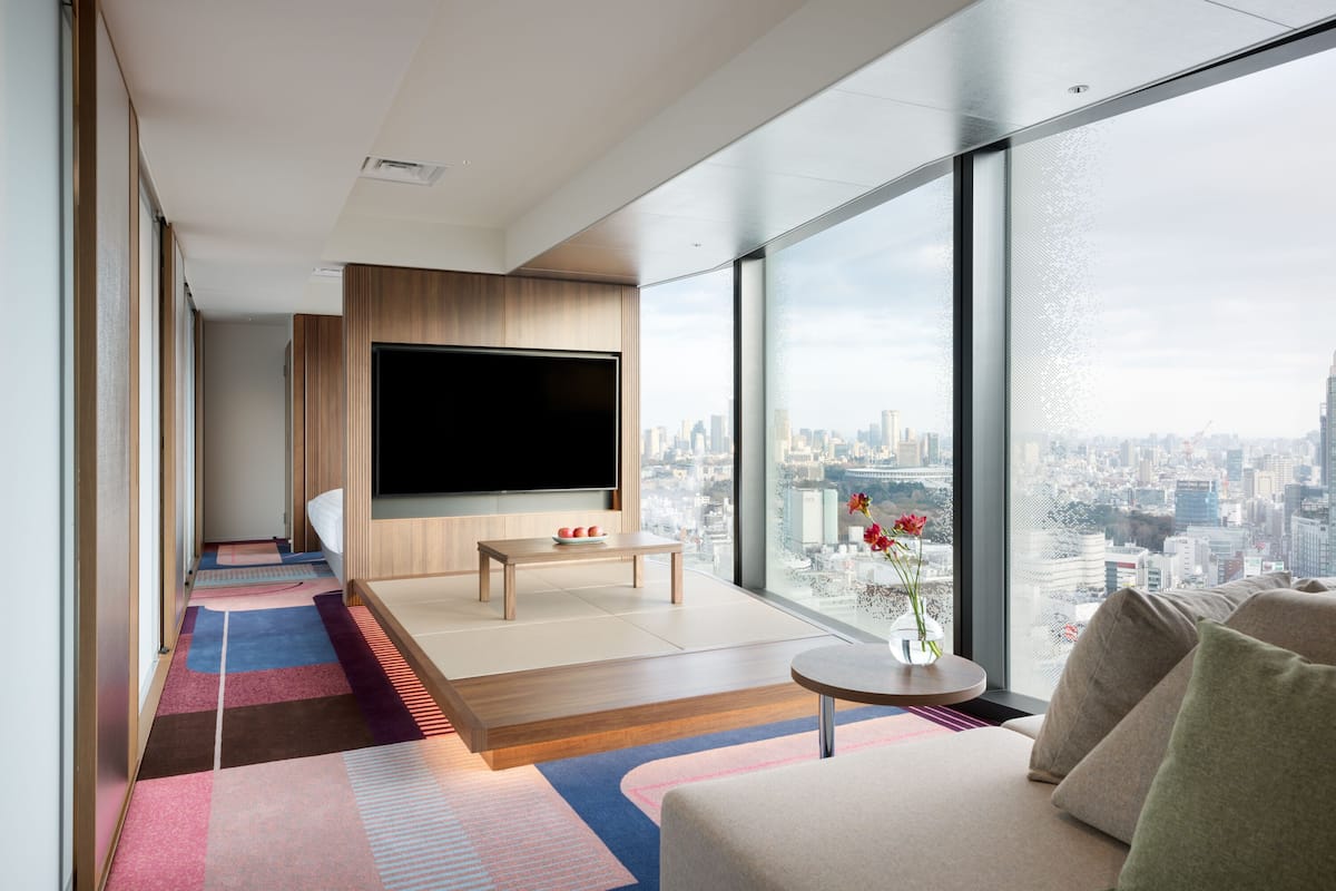 Ett modernt hotellrum med en stor TV, en soffa, ett litet bord och utsikt över staden.