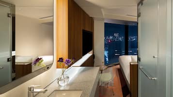 A modern hotel bathroom with a large mirror, a sink, a toilet, and a shower. There is a view of the city at night through the window.