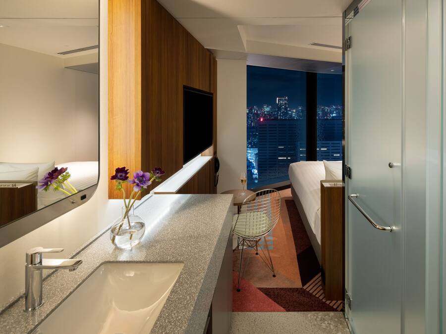 A modern hotel bathroom with a large mirror, a sink, a toilet, and a shower. There is a view of the city at night through the window.