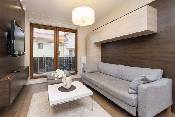 The living room features a modern gray sofa, a coffee table, and large windows that let in natural light. The space is cozy yet contemporary, with neutral tones and wooden flooring.