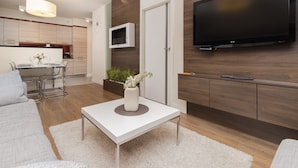 Una sala de estar moderna con un sofá blanco, una mesa de centro con un florero, un televisor de pantalla plana montado en la pared y una cocina al fondo.