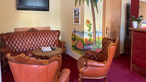 A room with a television mounted on the wall, a large painting of a palm tree, a wooden door, and a small table with a chair.