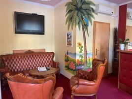 Quarto com televisão na parede, uma pintura grande de uma palmeira, uma porta de madeira e uma pequena mesa com uma cadeira.