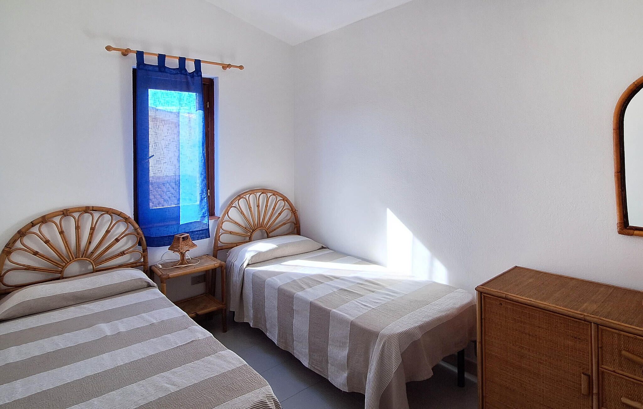 Une chambre avec deux lits simples, une table de chevet en bois avec une lampe, une fenêtre avec des rideaux bleus et un miroir au mur.