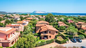 A coastal residential area with terracotta-roofed houses, a clear blue sky, and a distant mountain range.