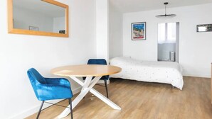 Ein kleiner, heller Raum mit Holzboden, einem runden Tisch mit zwei blauen Stühlen, einem Bett mit weißer Bettdecke und einem Spiegel an der Wand.