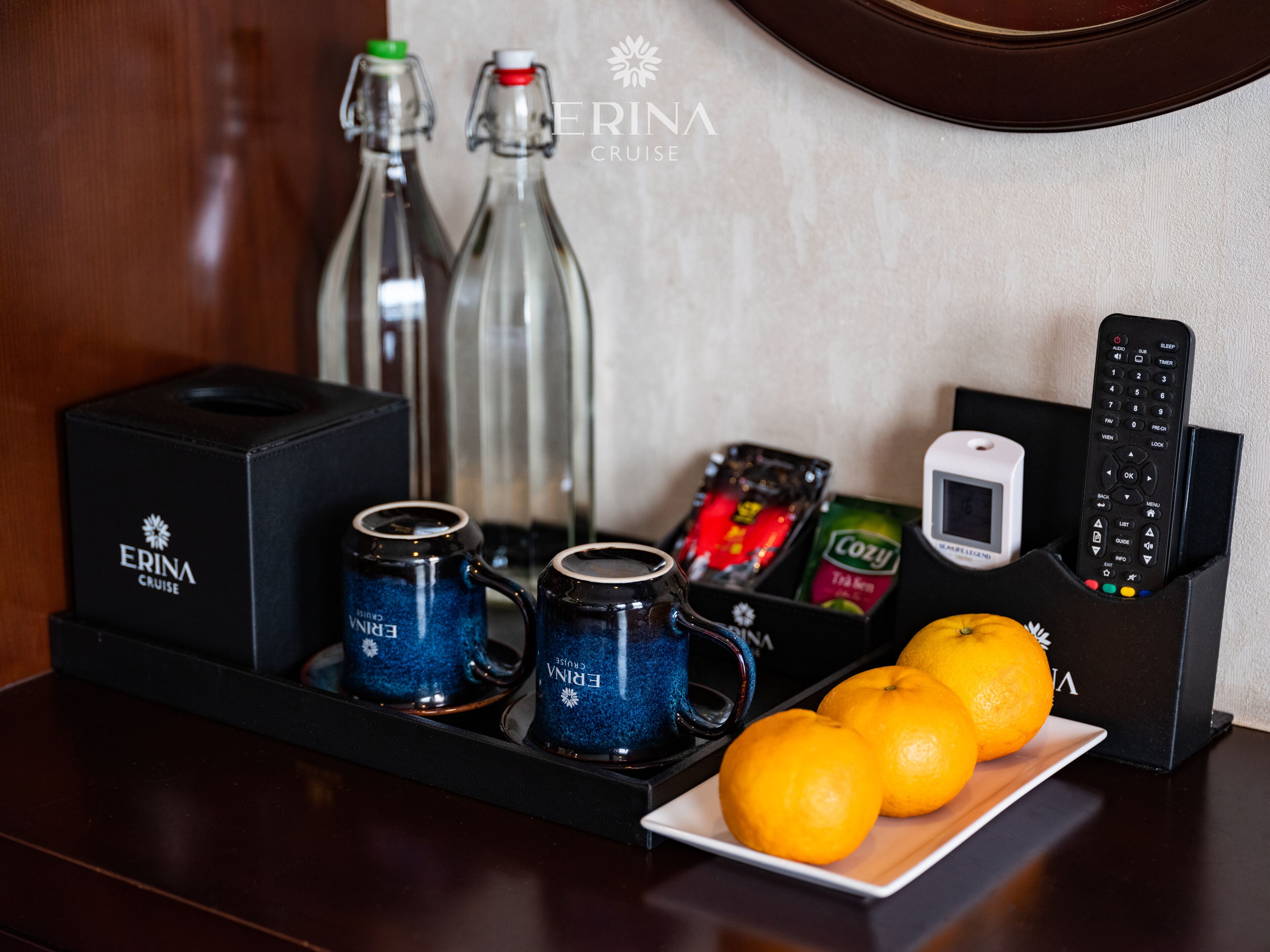 Una bandeja con dos tazas azules, dos botellas, una caja etiquetada 'ERINA CRUISE', un control remoto, una caja de té 'Cozy' y dos naranjas sobre una mesa.