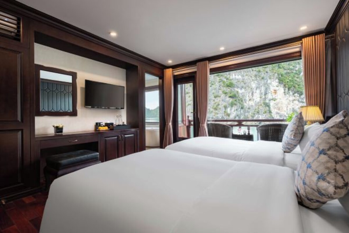 A hotel room with two beds, a desk, a TV, and a balcony with a view of greenery.