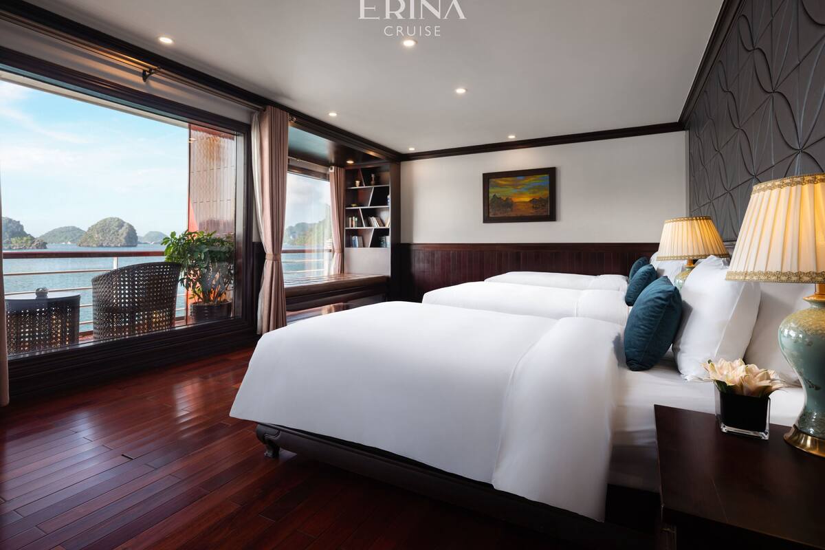 A modern hotel room with a large bed, a view of the sea, a bookshelf, and a painting on the wall.