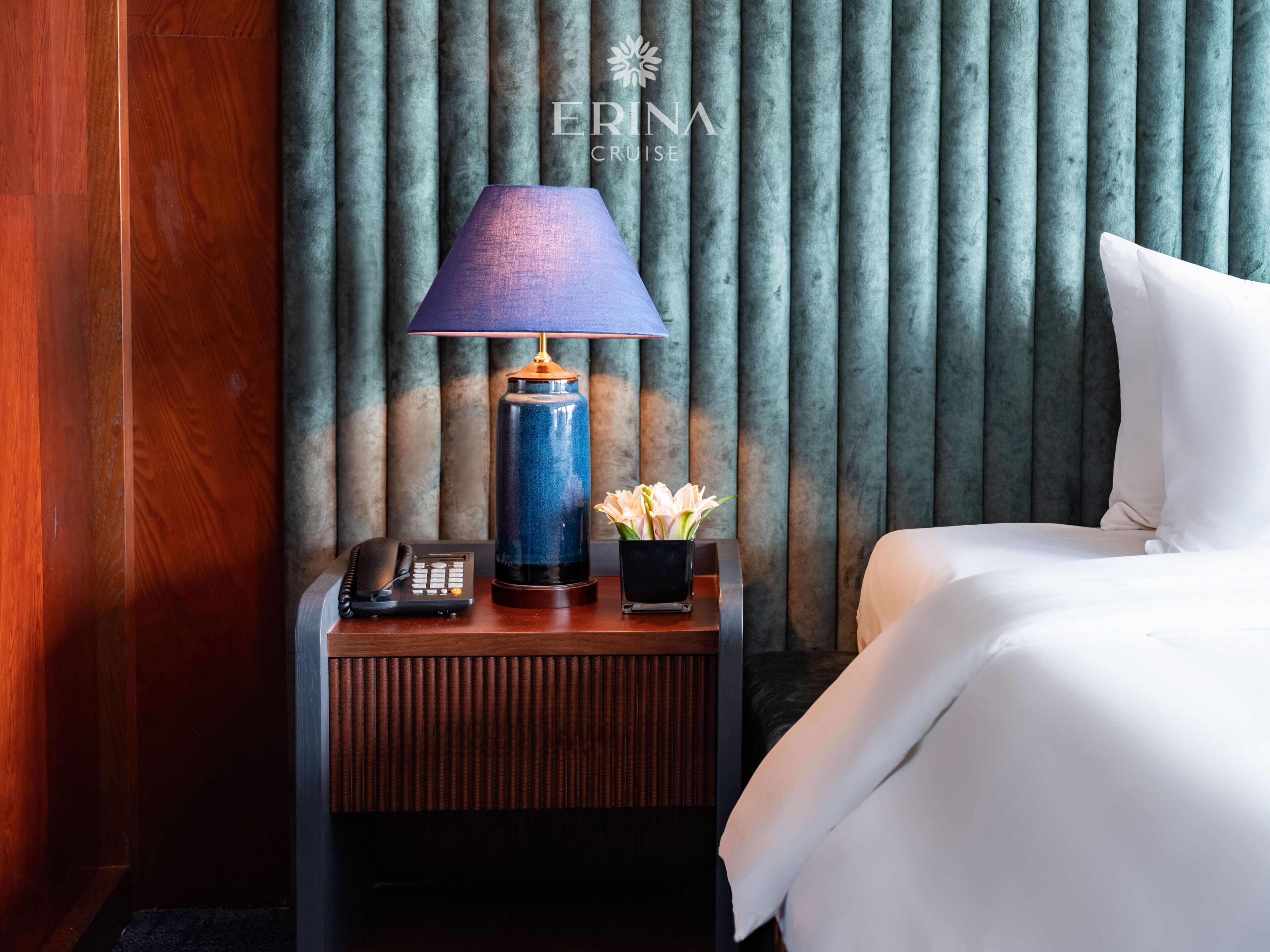 A hotel room with a wooden nightstand, a lamp, a vase with flowers, a phone, and a bed with white bedding.