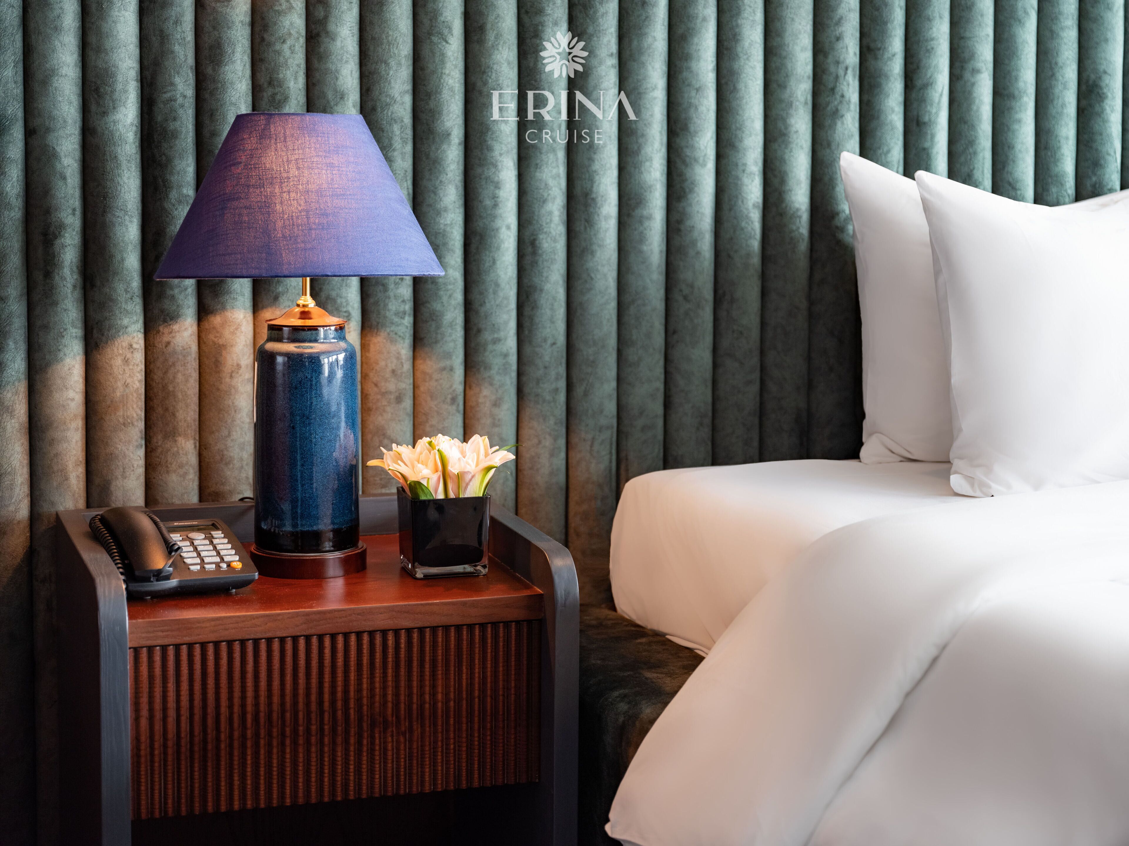 A hotel room with a bed, a nightstand with a lamp, a telephone, and a vase with flowers.
