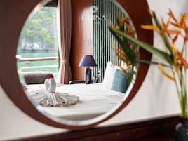 A hotel room with a bed, a lamp, a vase with flowers, and a view of greenery outside.