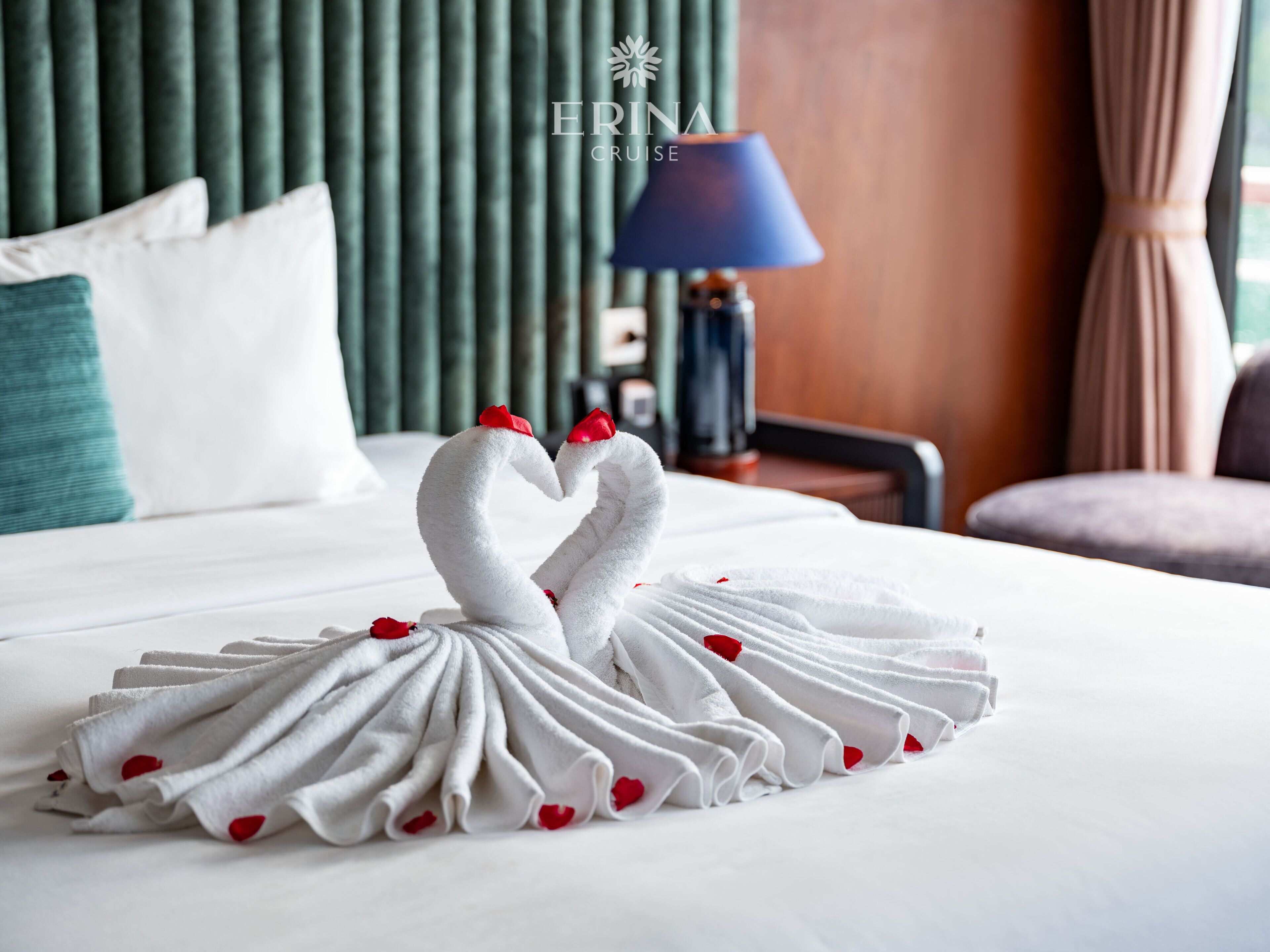 A neatly made bed with a towel swan centerpiece, a blue lamp, and a green headboard.