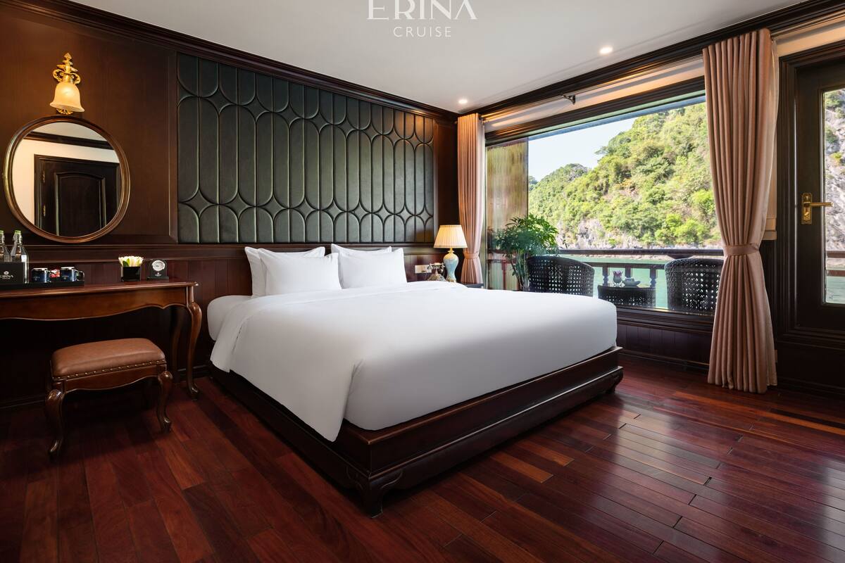 A spacious hotel room with a large bed, a wooden desk, a mirror, and a view of greenery outside.