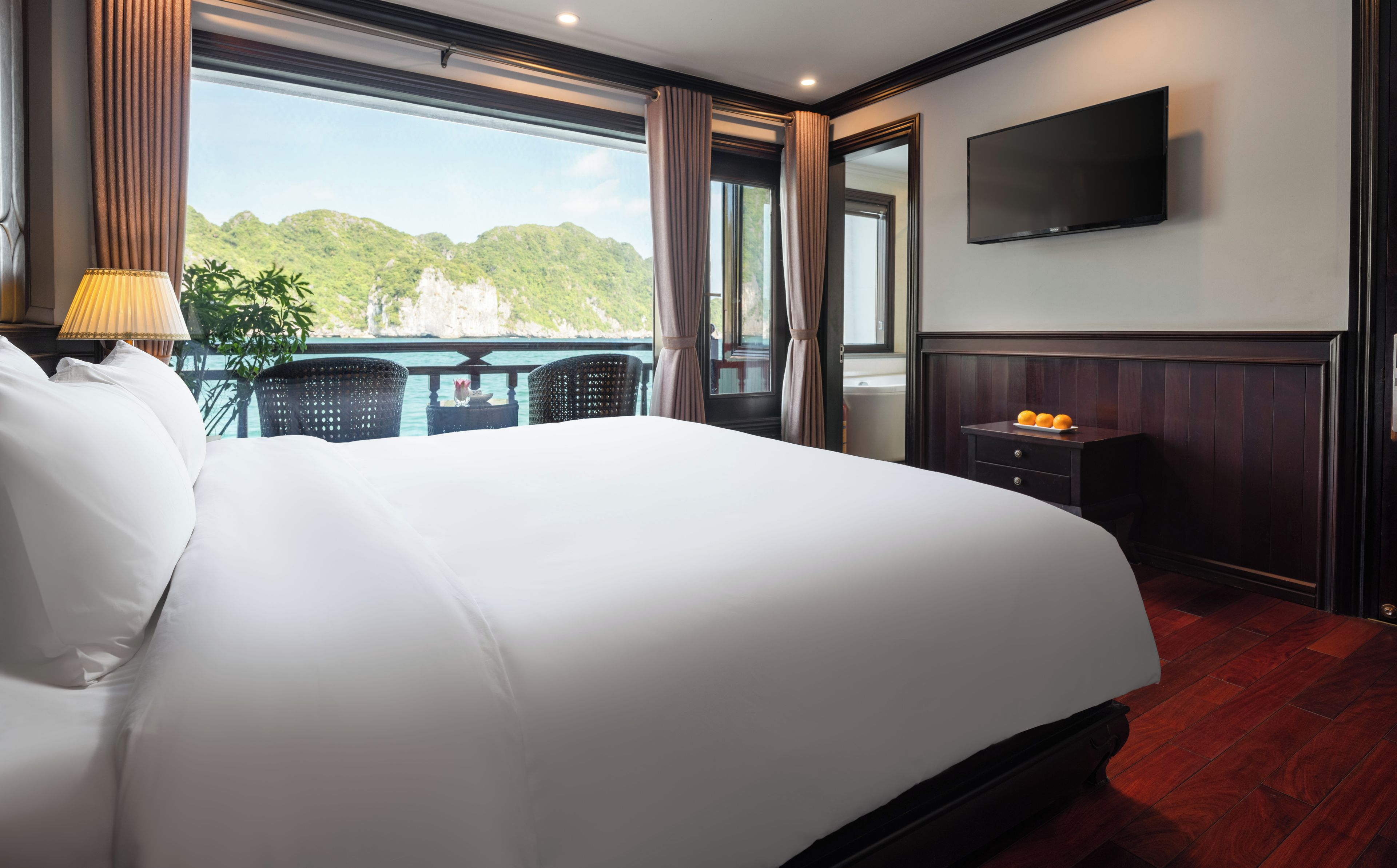 Quarto de hotel com uma cama grande, televisão, varanda com vista para o mar e uma mesa com laranjas.