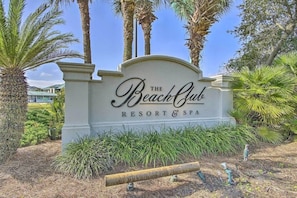 A sign for The Beach Club Resort & Spa surrounded by palm trees and greenery.