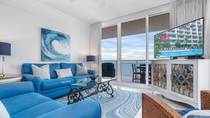 Een moderne woonkamer met een blauwe zithoek, een glazen salontafel en een groot televisiescherm waarop een uitzicht op een hotel te zien is.