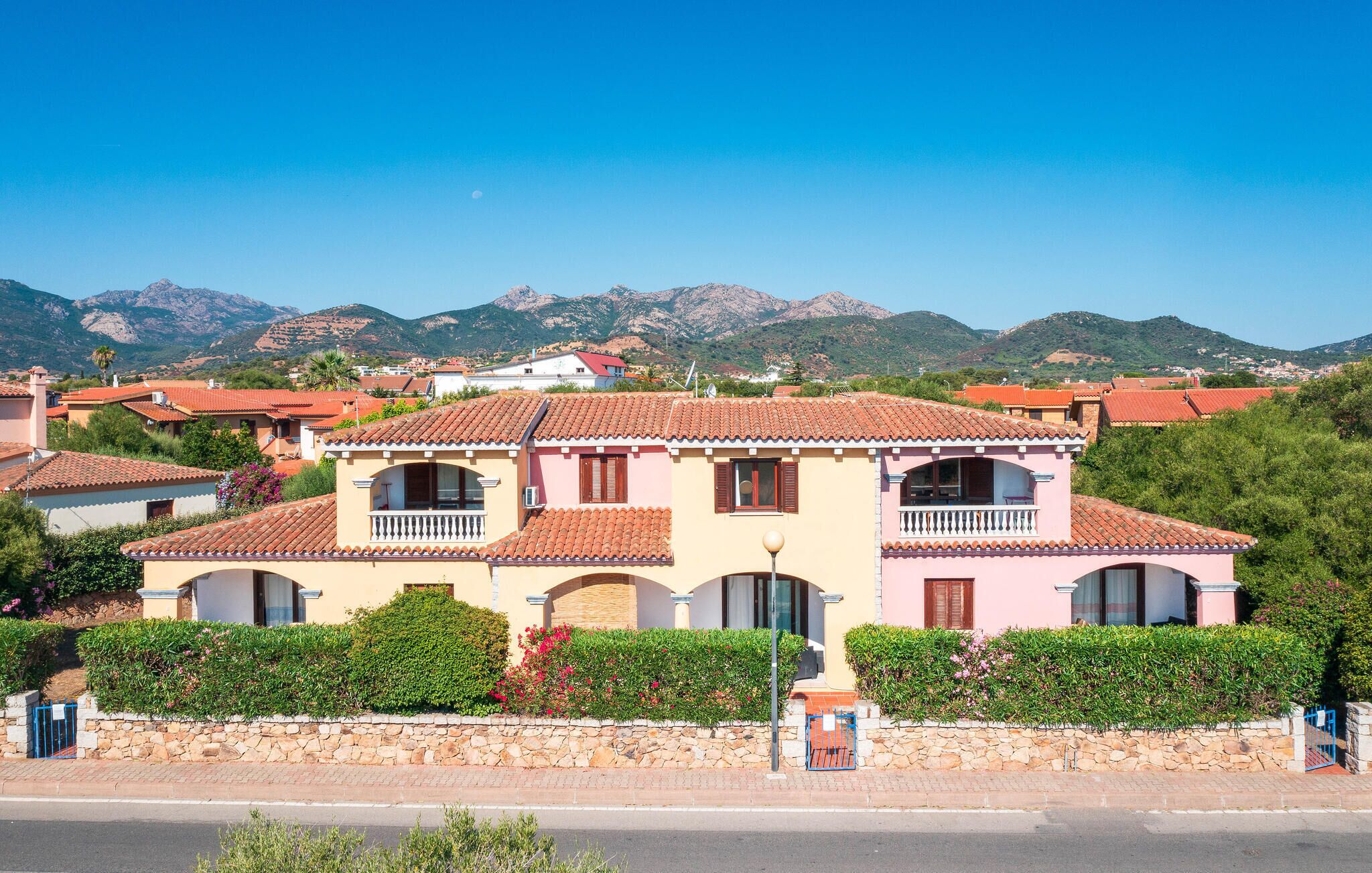 A two-story house with a tiled roof, a stone wall in front, and a mountain backdrop.