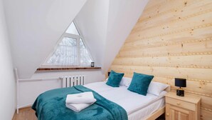 Een slaapkamer met een schuine kap, een houten hoofdbord, een bed met blauwgroene beddengoed, een nachtkastje met een lamp, en een raam met uitzicht op bomen.