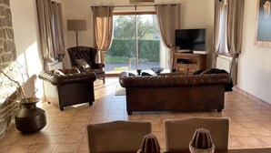 A spacious living room with a large window, a leather sofa, a television, and a dining table with candles. - Light & airy single-storey house with large terrace & heated outdoor pool (Isigny-le-buat)