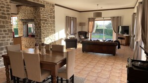 A spacious living room with a stone wall, a dining area with a wooden table and chairs, a leather sofa, and a television. - Light & airy single-storey house with large terrace & heated outdoor pool (Isigny-le-buat)