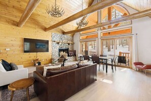 A spacious living area with a stone fireplace, wooden ceiling beams, and a large window overlooking a snowy landscape.