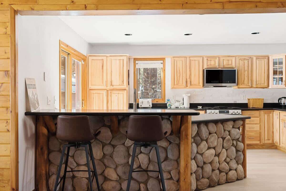 A kitchen with wooden cabinets, a stone island, two bar stools, a television, and a window.