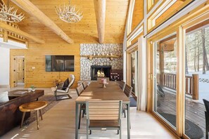 A spacious living area with a fireplace, wooden ceiling beams, and a large window overlooking a deck.