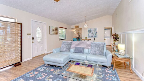 A living room with a light blue sectional sofa, a glass coffee table, a ceiling fan, a dining area with a table and chairs, and a kitchen in the background. - Cozy 1BR Retreat w/ Porch – Near Beaches & Airport (Bradenton)